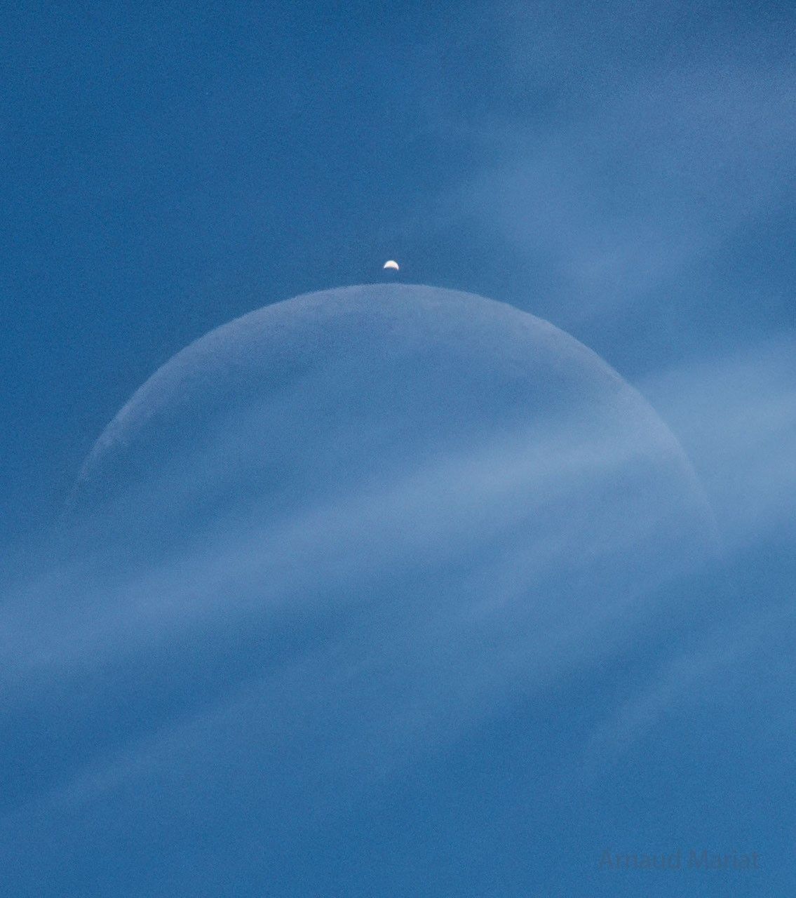 A blue sky has a light cloud crossing the middle of
the image. Two crescent objects are visible through the
cloud, a large faint crescent in the center and a brighter
crescent above it.
Please see the explanation for more detailed information.